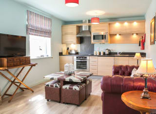 An open-plan view of one of our apartments. A wooden modern kitchen spreads across the back wall, with a red leather sofa on the right side, a compact four-seater table in the middle with a TV opposite the sofa. image