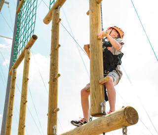 A pre-teen child is climbing on a wodden structure with a bright orange helmet, torso harness and a pully rope system. The child is looking up to judge his next move as he accends higher on the structure.