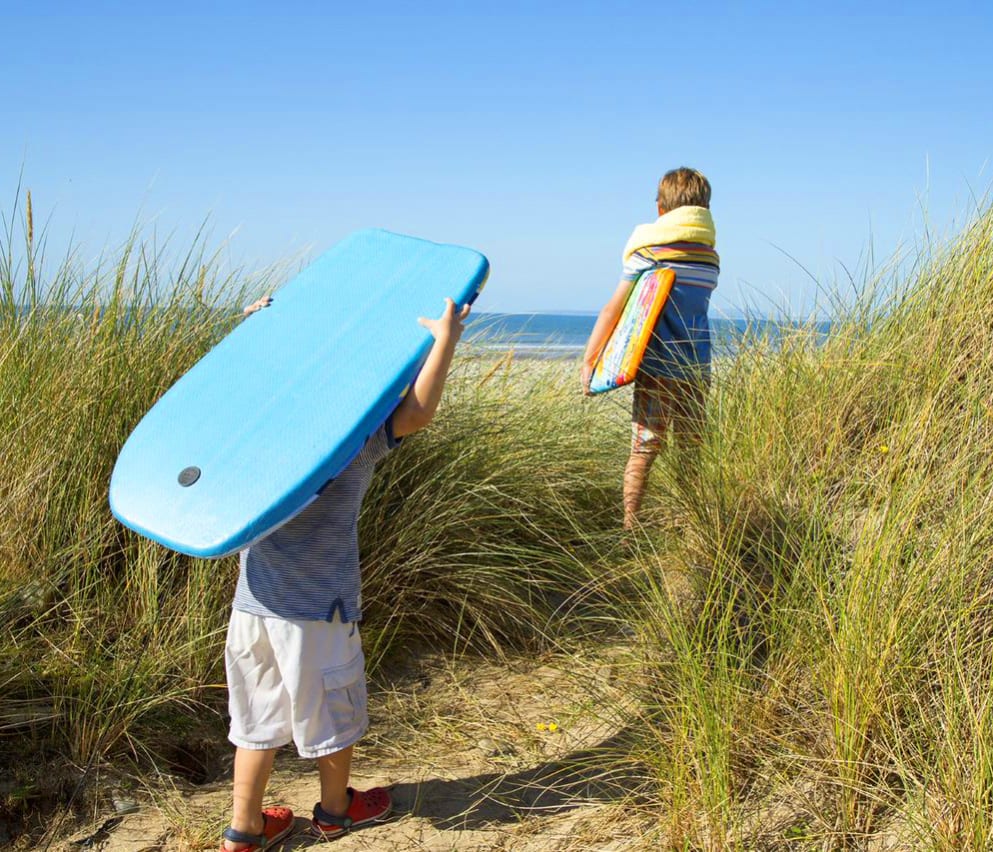 Two children walking across the sand to the sea holiday two body boards on a sunny day.