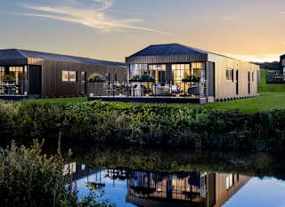 One of our holiday home barns. The sun is setting behind the spacious barn exterior, which sits on the green grass near the lake. The lights are on inside. image