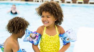 Two young children paddling in an outdoor pool. One is wearing a bright yellow swimming costume with armbands and the other is mostly out of shot but some of their curly hair is in frame.
