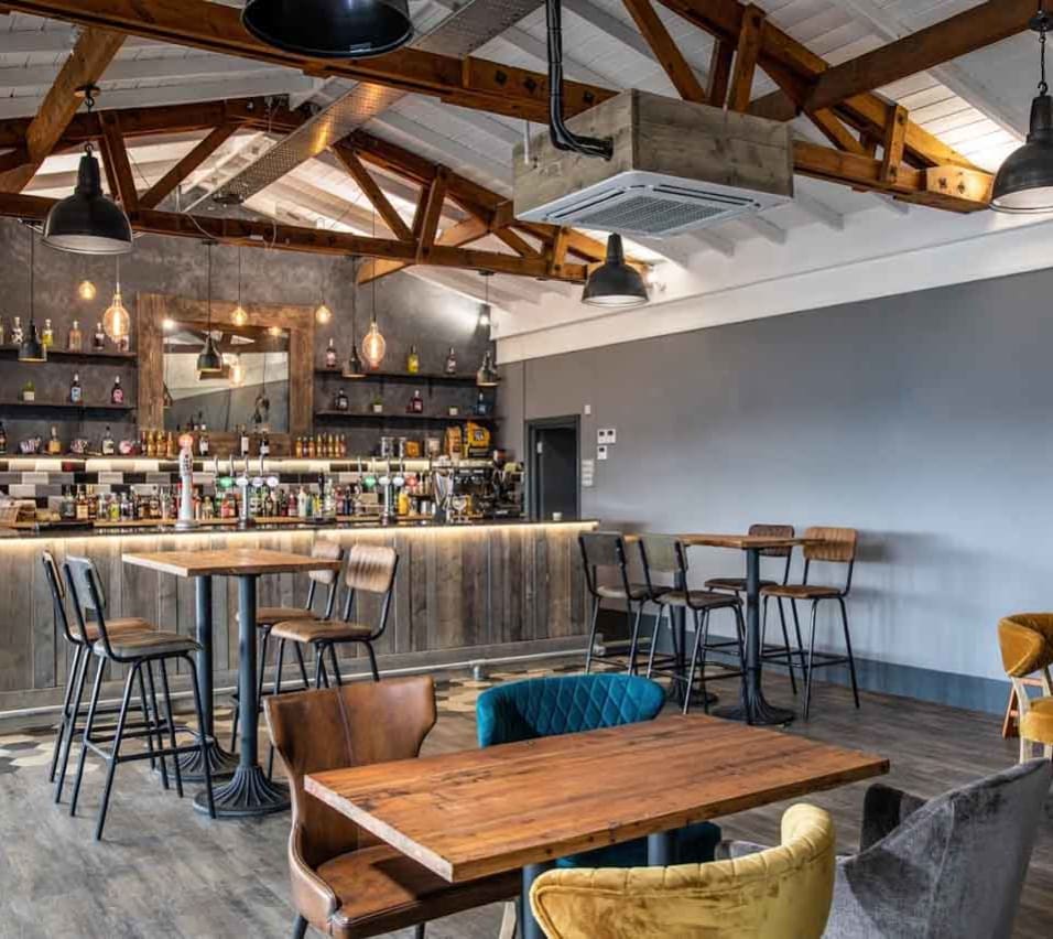A restaurant bar area, with grey walls and wood panelling. Four tables with chairs can be seen in the image. The bar is stocked with bottles on the back wall with a mirror hanging above. image