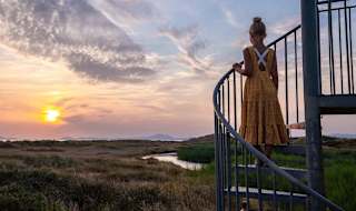 A photo of a girl with blonde hair in a bun wearing a orange polkadot dress, whom is standing in the middle of a spiral staircase looking out at the sunset.