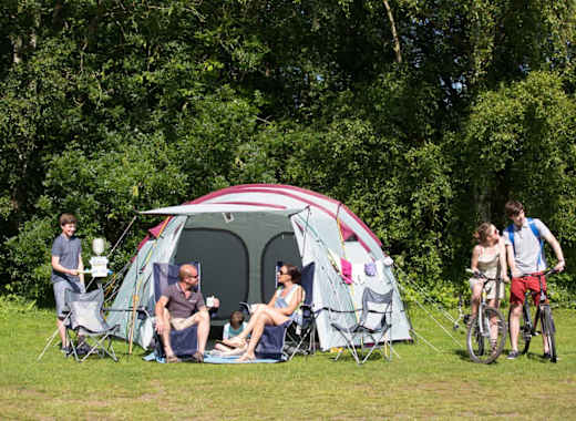 A pitched up family tent with a man, woman and baby sitting in the porch area of the tent. A teen boy and girl are cycling by on a hot sunny day.