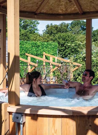A couple enjoying a break in their hot-tub at one of our holiday parks. The man has short black hair, and the woman has long brown hair in a bun. The man is wearing glasses, and they're both sitting in the shaded side of the hot tub.