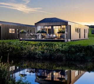 One of our holiday home barns. The sun is setting behind the spacious barn exterior, which sits on the green grass near the lake. The lights are on inside. image