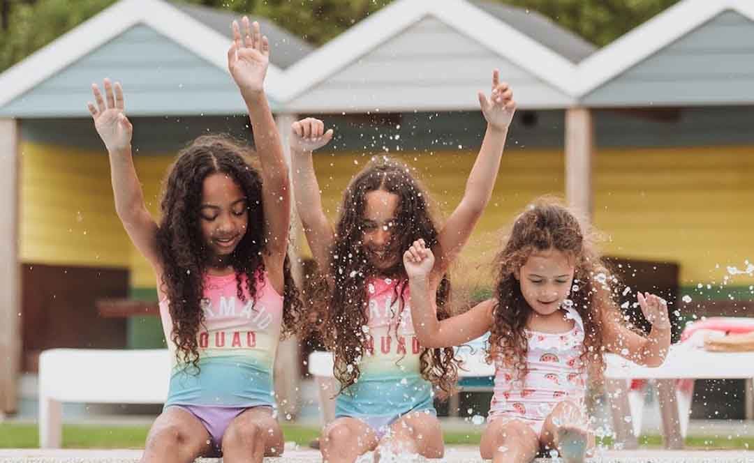 Three girls, each with long brown curly hair and pink swimsuits, are splashing and paddaling on the edge of our outdoor pool at Newquay Bay, Cornwall image