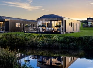 One of our holiday home barns. The sun is setting behind the spacious barn exterior, which sits on the green grass near the lake. The lights are on inside. image