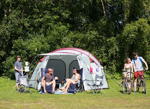 A pitched up family tent with a man, woman and baby sitting in the porch area of the tent. A teen boy and girl are cycling by on a hot sunny day.