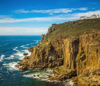 A landscape image of the sea and cliffs located at Lands End. A blue sky with minimal clouds sets the scene with the Land's end tourist building seen in the top right of the image.