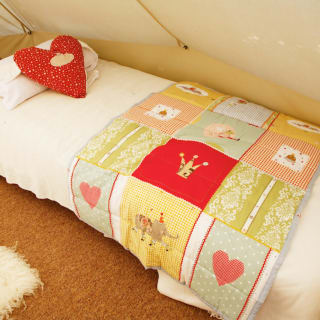 A photo of the bedroom area within a off-white canvas bell tent. The bed is a single camping bed with a cream duvet, pillow and red spotted heart-shaped cushion.