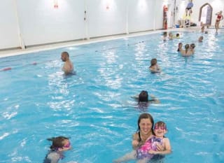 Lots of people of all age ranges swimming in an indoor swimming pool. There is a lifeguard sitting at the site of the pool on a lookout chair. image
