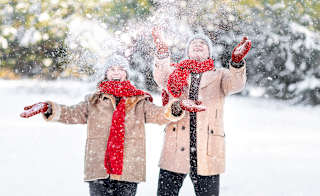 A man and a woman in light brown winter coats, red gloves and scarfs and grey benie hats are throwing snow up in the air and laughing as the snow crashes down on them. image