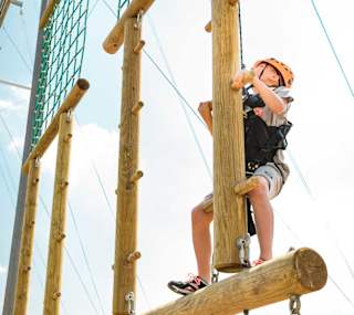 A pre-teen child is climbing on a wodden structure with a bright orange helmet, torso harness and a pully rope system. The child is looking up to judge his next move as he accends higher on the structure. image