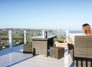 A woman sat on outdoor furniture on the decking of one of our stunning holiday homes at Swanage Bay View reading a book. There is a bottle of wine and a wine glass on the table and views of the surrounding areas and the sea can be seen in the distance. image