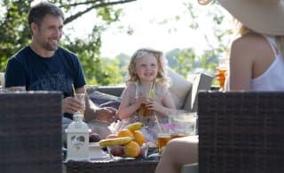 A family with a young child sitting outside enjoying a drink. On the table is a bowl of oranges and bananas. image