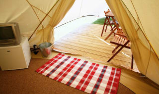 A photo of the living space inside of a canvas bell tent. To the left of the photo is a silver TV propped up on a white box. In the background is the entrance to the bell tent. image