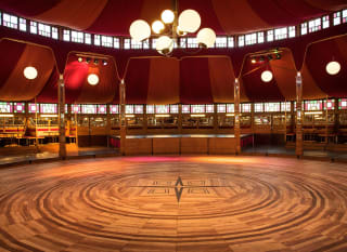 A photo of a large circular wooden dancefloor in the middle of a huge tipe-style tent. There are sphare-shaped lights hanging off the wooden support pillars for decoration. The tent roof is coloured red and yellow split. image