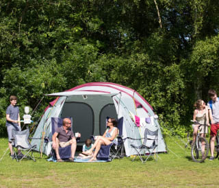 A pitched up family tent with a man, woman and baby sitting in the porch area of the tent. A teen boy and girl are cycling by on a hot sunny day.