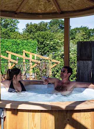 A couple enjoying a break in their hot-tub at one of our holiday parks. The man has short black hair, and the woman has long brown hair in a bun. The man is wearing glasses, and they're both sitting in the shaded side of the hot tub.