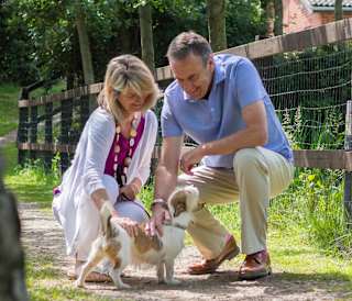 A mature man and woman are petting a light brown and white fluffy dog. Both the man and woman are kneeling down and smiling.