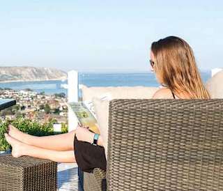 A woman sat on outdoor furniture on the decking of one of our stunning holiday homes at Swanage Bay View reading a book. There is a bottle of wine and a wine glass on the table and views of the surrounding areas and the sea can be seen in the distance.