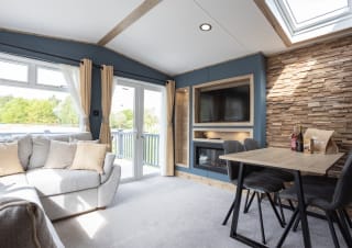 The inside of one of our static accessible caravans. There is a grey sofa and a dining area, along with a TV and fireplace. The large double doors are letting in sunlight.