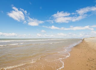 A landscape view of a sandy stoney beach and the sea. The waves are rolling into shore. The sky above is soft blue with multiple clouds. image