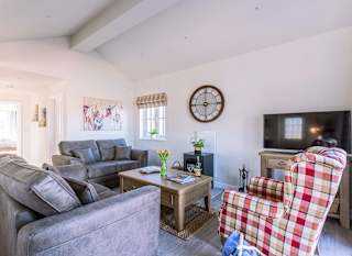 An interior to a holiday lodge for sale. The living room has two grey sofas, a red checked armchair, a wall mounted clock and a flat screen TV. image