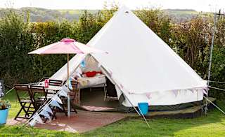An off white canvas bell tent pitched up outside on the grass on a sunny day. The entrance to the bell tent is open and there is a pink parasol resting against the entrance. image