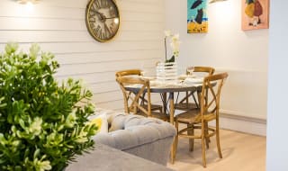 A glimpse of the dining area in one of our cottages. A four-seater round table set ready for dinner. two pictures and a clock hanging on the walls around the table. A section of a sofa is seen in the left hand corner. image