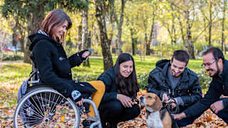 two women and two men are pictured with a medium sized dog. One woman is in a wheelchair and the others are crouched down petting the dog.