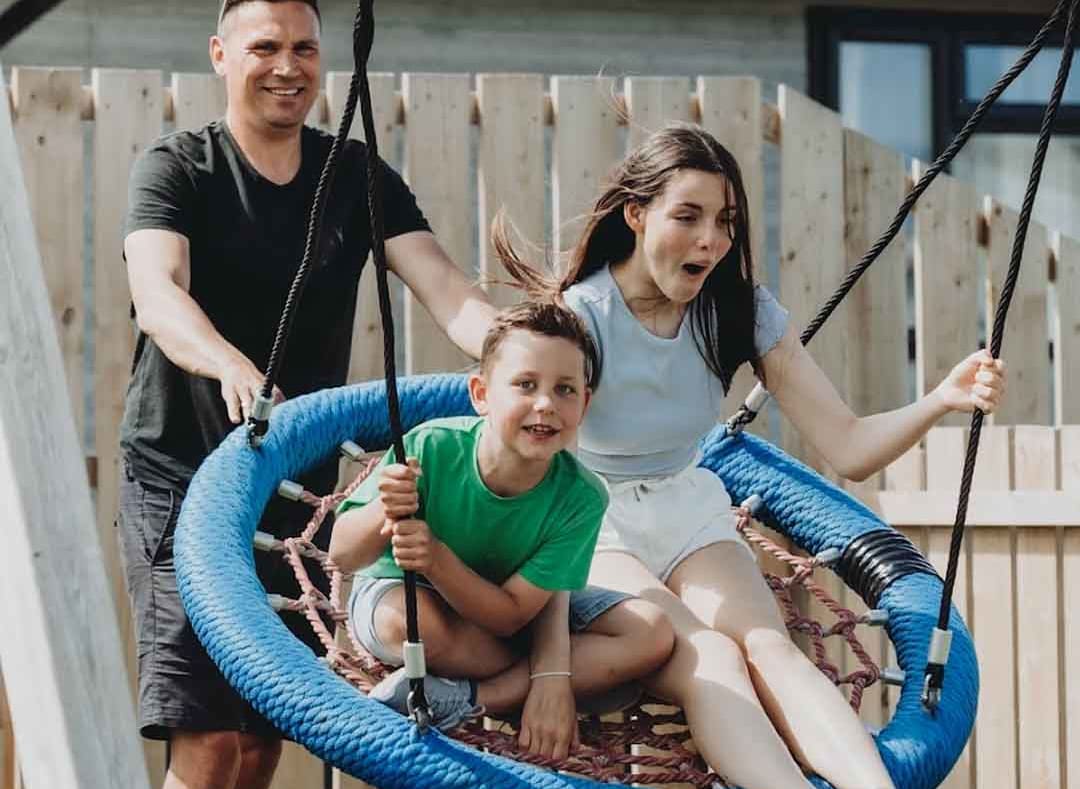 Two children playing on the swing at our adventure playground at Bude Holiday Park. There is a male pushing the swing. image
