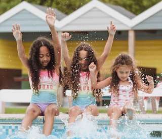 Three girls, each with long brown curly hair and pink swimsuits, are splashing and paddaling on the edge of our outdoor pool at Newquay Bay, Cornwall