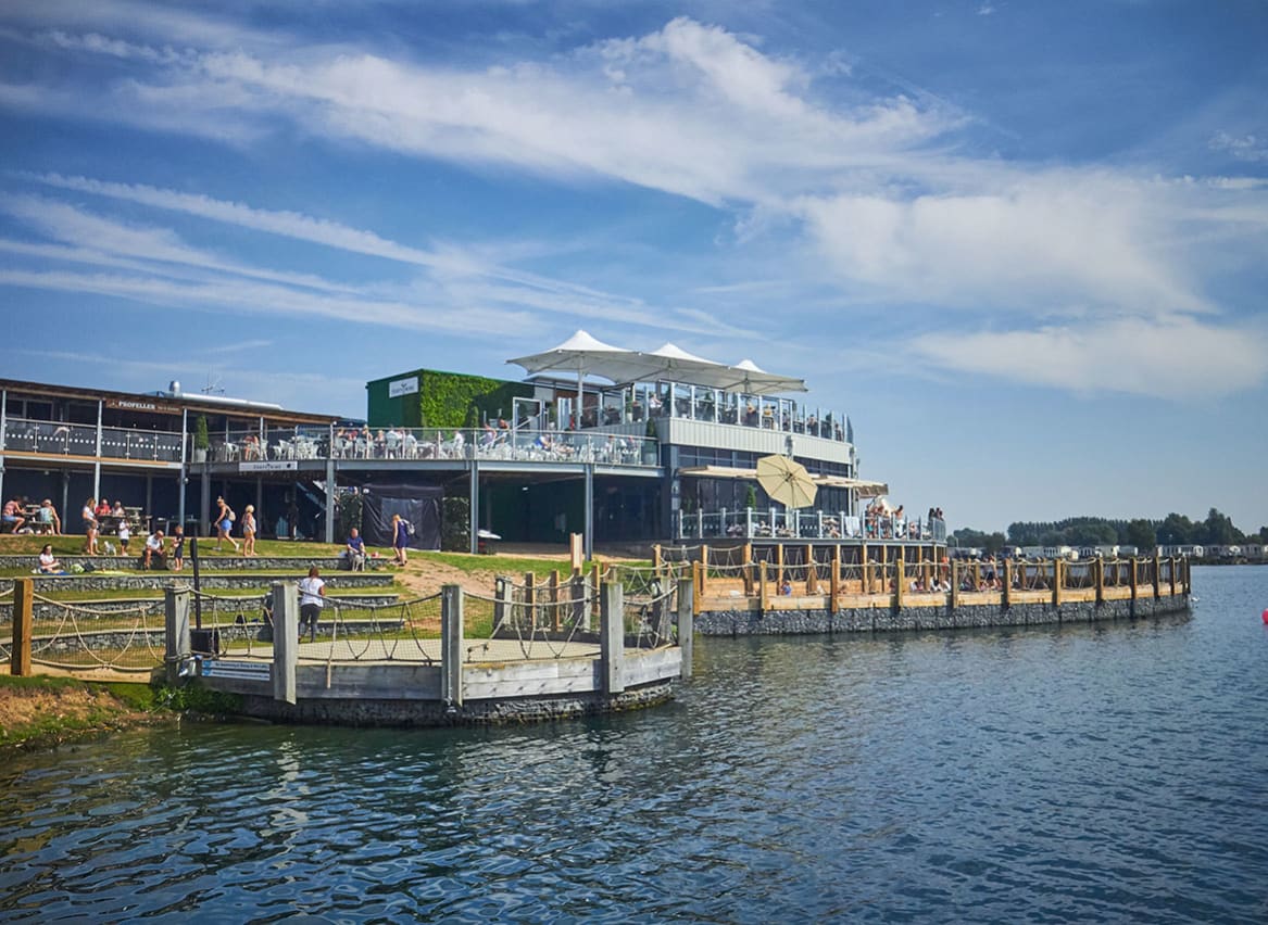 A photo of a restaraunt with a ground floor and two balconey levels of outdoor steating. There are lots of people sitting across all levels and the building is next to a large river. image