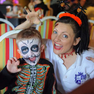A young white boy with a skeleton face paint and outfit is holding his hand up like a claw with a white woman who is wearing face paint-style injuries to her head and neck, whilst also showing both her hands like claws. image