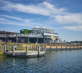 A photo of a restaraunt with a ground floor and two balconey levels of outdoor steating. There are lots of people sitting across all levels and the building is next to a large river. image