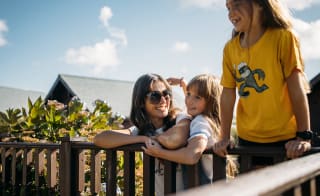 A family enjoying their stay at one of our holiday parks. There is an adult woman and two young girls. One of the girls is hugging the woman, the other is leaning over a banister. All three have wide smiles. image