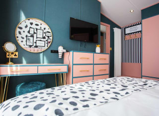 In the double bedroom is a large light pink and gold vanity table with a large circular mirror on the wall and a flat screen TV hanging on the wall next to it. image