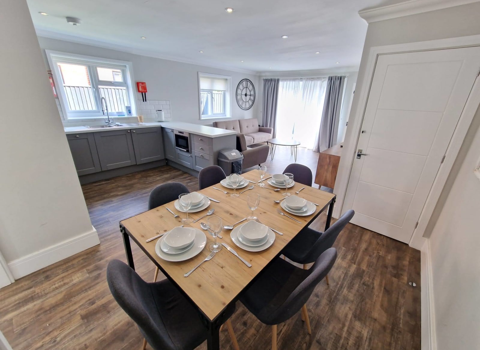 An open-plan view of one of our cottages. In front view is a dinner table set for six. across from the table is a U-shaped Kitchen area with plenty of worktop space. In the background is the living room area containing two sofas. image