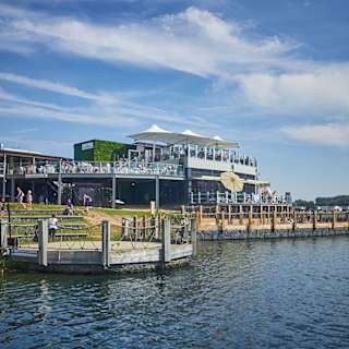 A photo of a restaraunt with a ground floor and two balconey levels of outdoor steating. There are lots of people sitting across all levels and the building is next to a large river.