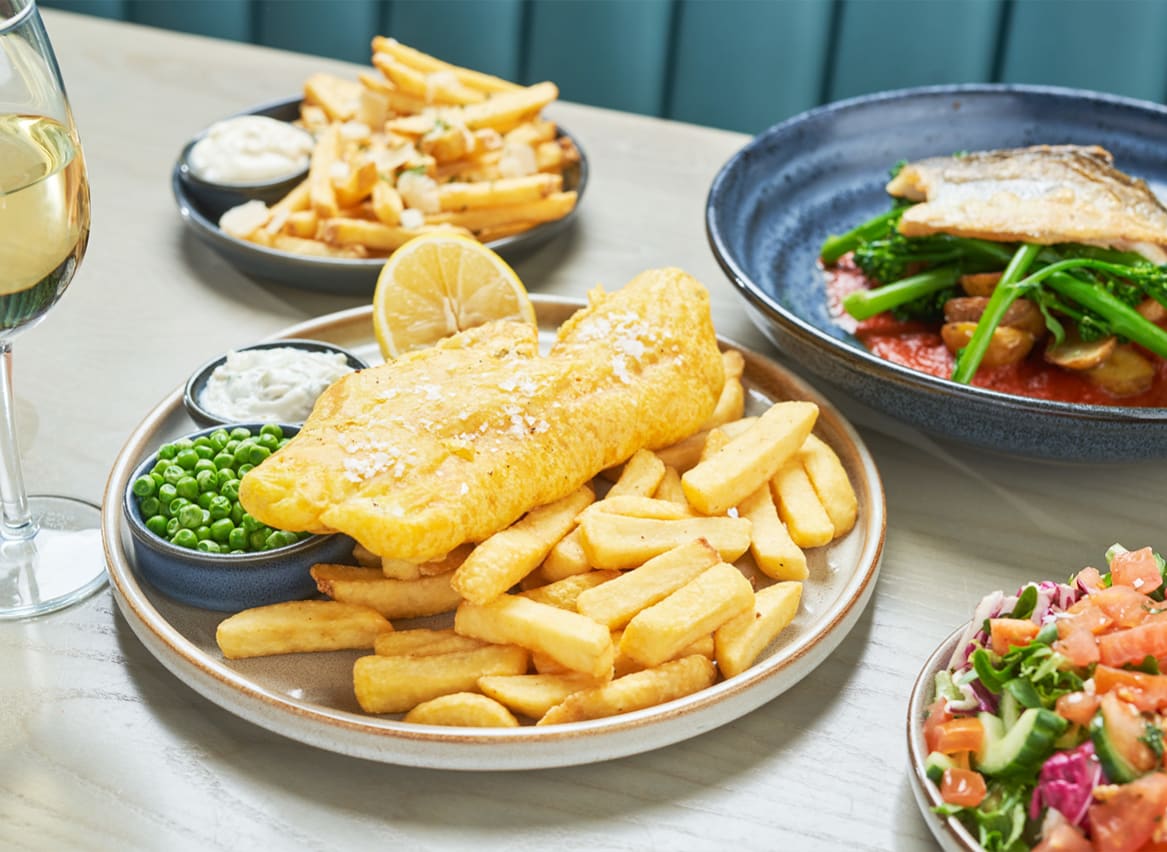 A close up of fish and chips on a wooden table with various other dishes around it. image