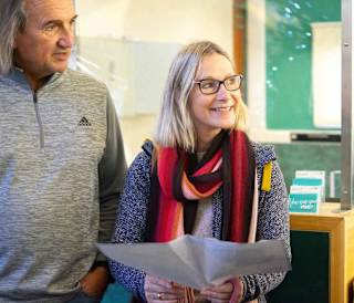 A photo of a mature man and woman mid sentence. The man has long grey hair and is wearing a grey pullover jumper. The woman also has long grey hair and is wearing a scarf, glasses and a dark blue hoodie. She is holding a map.