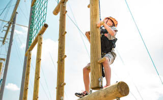A pre-teen child is climbing on a wodden structure with a bright orange helmet, torso harness and a pully rope system. The child is looking up to judge his next move as he accends higher on the structure. image