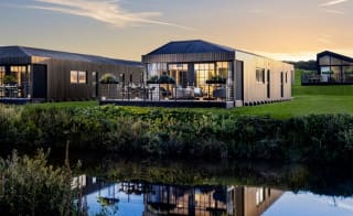 One of our holiday home barns. The sun is setting behind the spacious barn exterior, which sits on the green grass near the lake. The lights are on inside. image