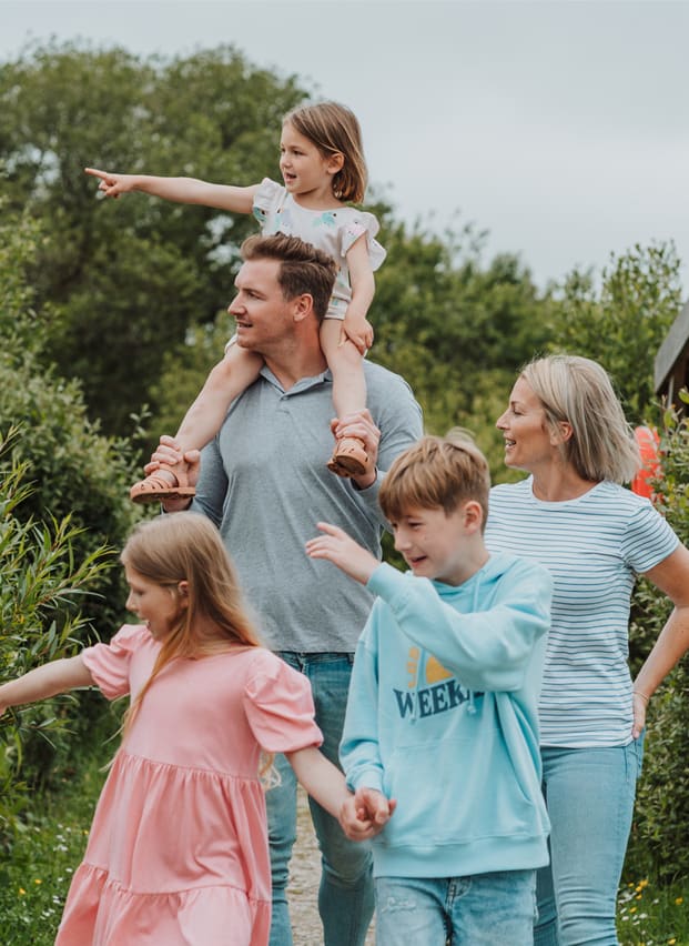 A white family of five, walk along a pathway. The youngest child, a girl, is sat on top of her father's shoulders. All five members of the family seem to be looking and pointing to something in the distance.