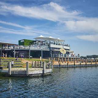 A photo of a restaraunt with a ground floor and two balconey levels of outdoor steating. There are lots of people sitting across all levels and the building is next to a large river.