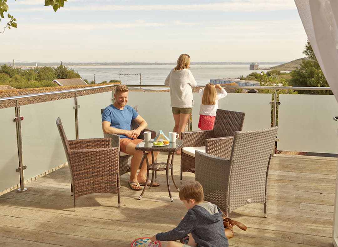 A family of four on the balcony of their holiday home in The Bay Colwell. There are two adults, a young boy, and a young girl with strawberry blonde hair, and all dressed in summer attire. image