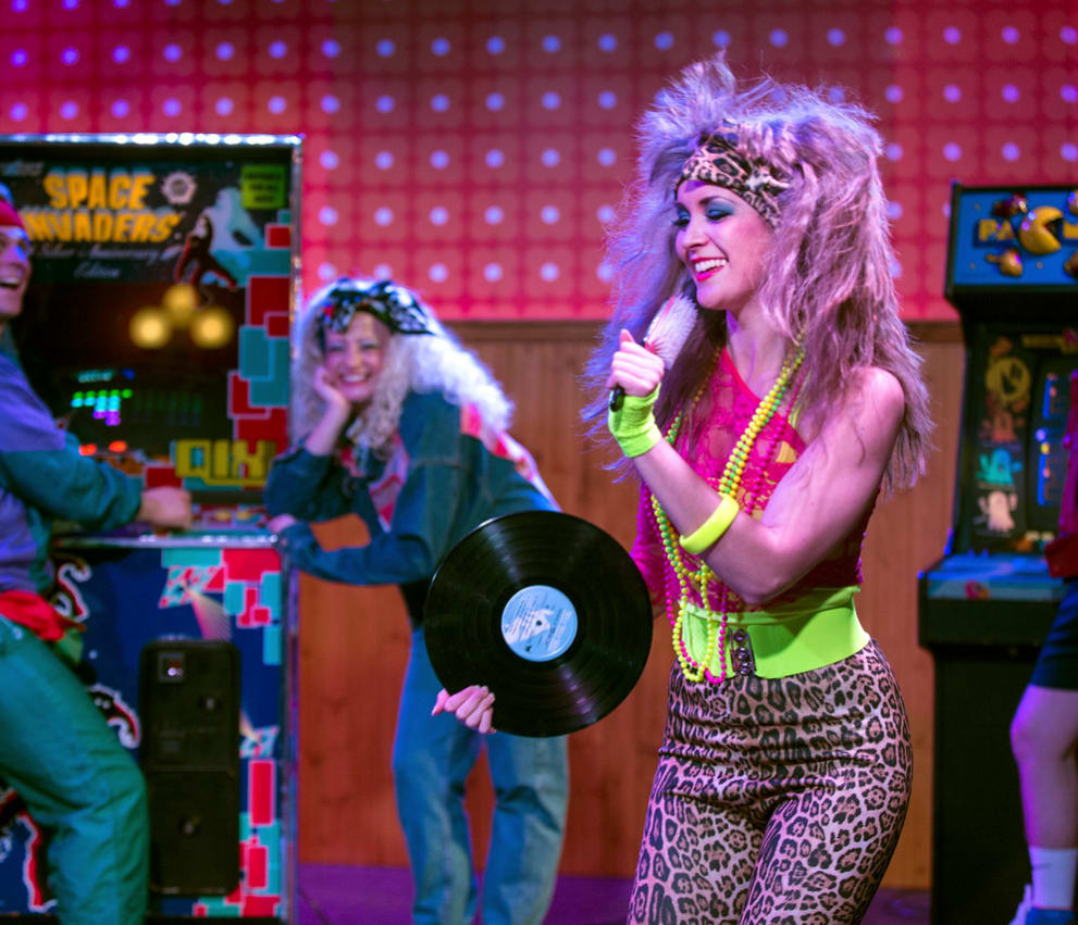 Two women are dressed up in 80's party attire with big curly hair inside an 80's themed disco with an arcade machine.