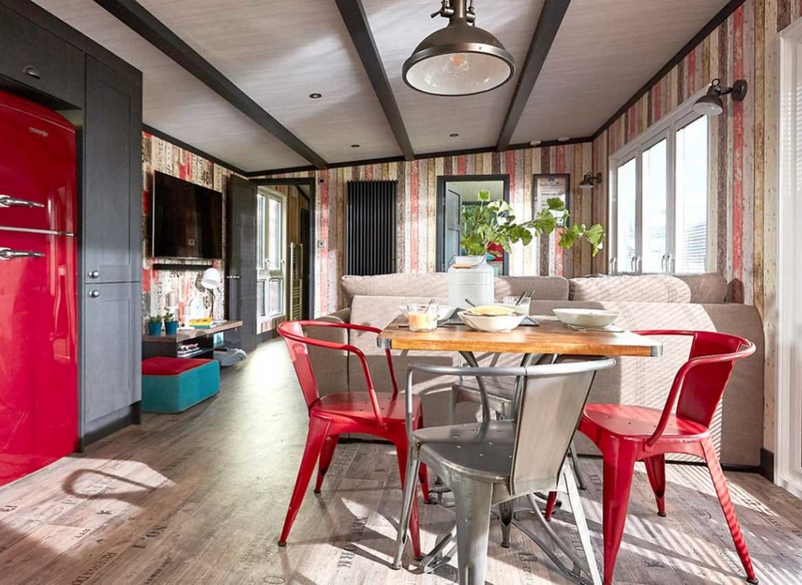 An image of the interior of one of our holiday lodges. There's wooden panneling, a cream fabric sofa and red metal dining chairs around a wooden table. image
